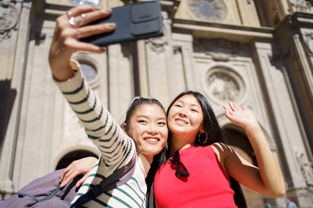 Delighted Asian women taking selfie on smartphone smiling during trip against Cathedral of Granada