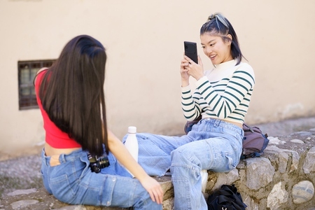 Smiling Asian woman taking picture of girlfriend on street