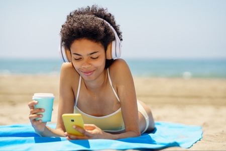 Happy black lady using smartphone and drinking takeaway beverage lying on seashore