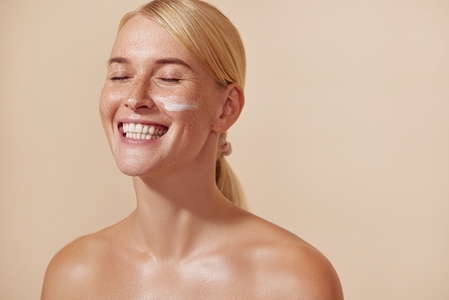 Happy woman with blonde hair and freckles  Young female with moisturizer on her cheek smiling with closed eyes in the studio