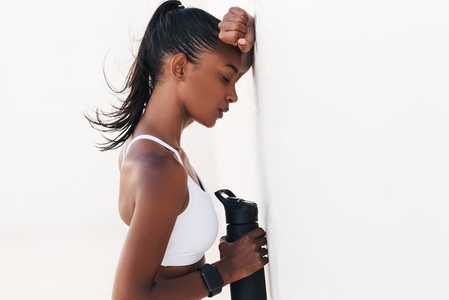 Side view of a tired and exhausted female with a bottle leaning her hand  Slim female with closed eyes taking a break during an intense workout standing with closed eyes