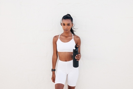 Slim woman in white fitness attire holding bottle leaning wall looking at the camera