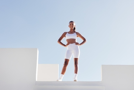 Slim smiling woman in white fitness attire posing on a top of stairs case  Full length fit female with hands on her hips looking at the camera while standing on a top