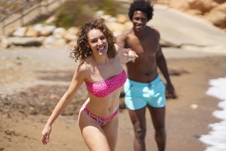 Cheerful diverse couple holding hands while running on beach