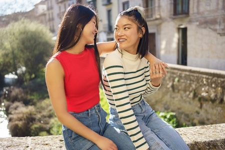 Happy Asian girlfriends sitting together on street and looking at each other