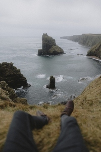 POV legs of man sitting on top of cliff at rugged coastline