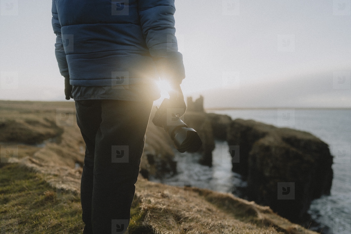 Photographer holding camera on cliff above ocean coastline at sunset
