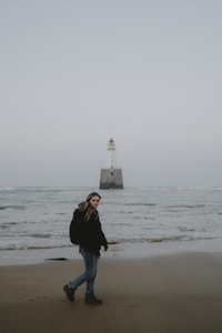 Portrait young woman walking past lighthouse on wet sand beach