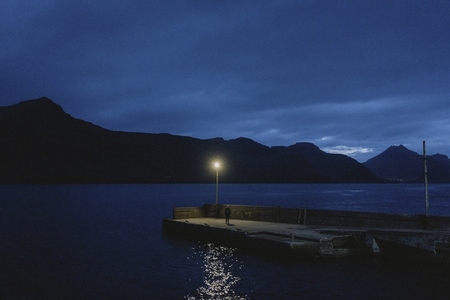 Man in dark on ocean jetty below streetlamp and dark blue night sky