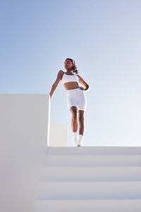 Full length of a young slim female posing while standing on the top of stairs  Woman in white fitness attire standing outdoors against the skies