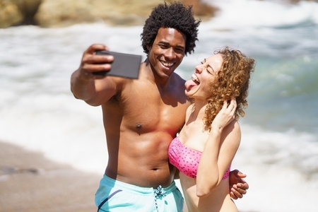 Cheerful diverse couple taking selfie on seashore during sunny day