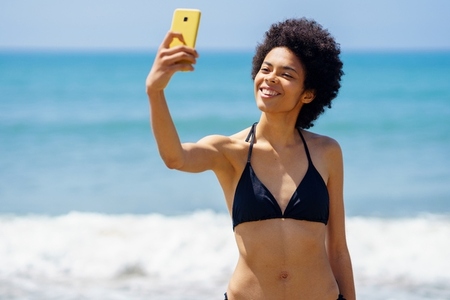 Gleeful black woman in swimwear taking selfie on seashore
