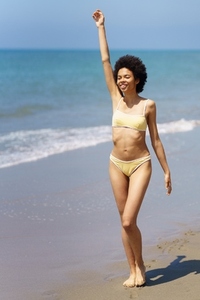 Happy ethnic woman with raised arm strolling on wet sandy seashore