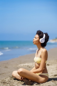 Calm black woman listening to music with closed eyes while relaxing on seashore