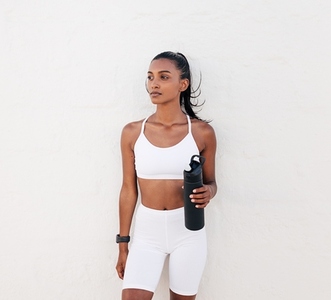 Confident fitness female athlete with bottle relaxing at a white wall
