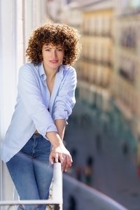 Curly haired woman in blue shirt and jeans leaning on metal railing of balcony