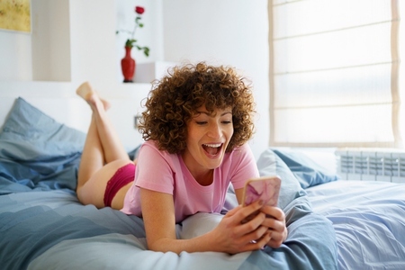 Excited woman using cellphone while lying in bed at home