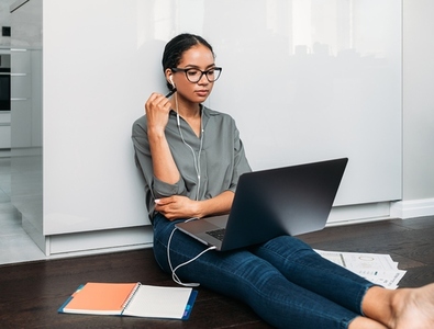 Young female entrepreneur in thought sitting on the floor in the kitchen with a laptop on her legs