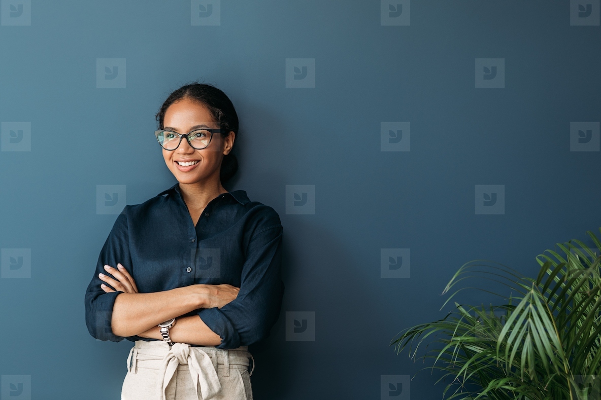 Confident businesswoman with crossed arms leaning on blue wall  Smiling entrepreneur in eyeglasses looking away