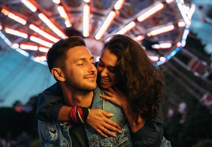 Smiling man piggybacks his happy girlfriend at the festival  Young couple having fun at night in an amusement park