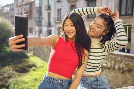 Joyful Asian female friends shooting selfie on bridge together