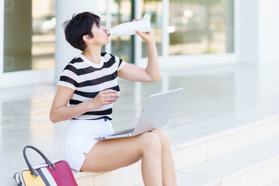 Calm woman drinking water and using laptop on stairs