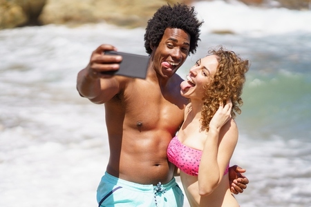 Funny multiethnic couple showing tongues while taking selfie on beach