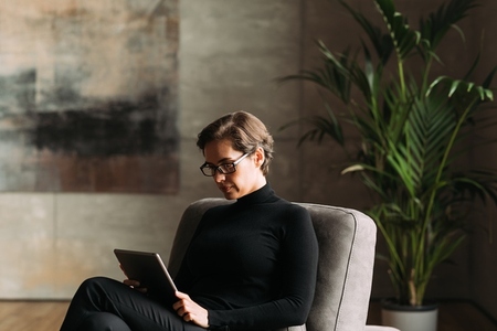 Middle aged businesswoman in black formal wear sitting indoors holding a digital tablet