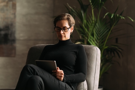 Serious adult woman in black formal wear sitting and reading from a digital tablet