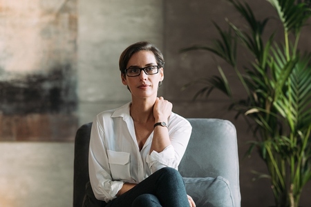 Confident businesswoman in eyeglasses sitting on a grey armchair in her loft