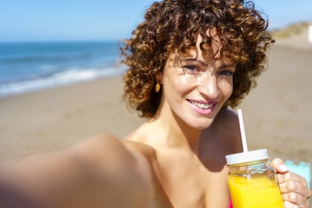 Cheerful woman enjoying juice and taking self portrait on beach