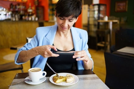 Smiling young woman sitting at table and taking photo with smartphone