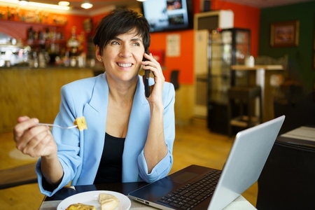Cheerful woman talking on smartphone while eating at table with laptop