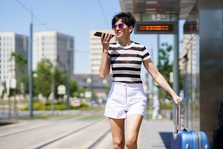 Stylish woman with suitcase recording voice message smartphone in city