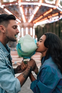 Side view of a young couple eating cotton candy against a carousel during the festival
