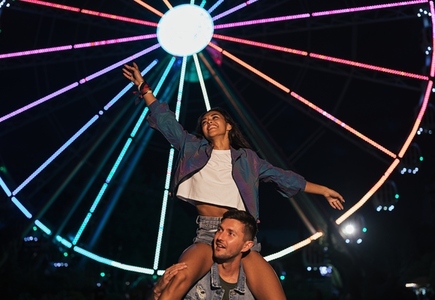 Happy woman sitting on the shoulders of her boyfriend and having fun  Young couple against ferris wheel during the festival