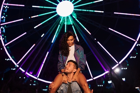 Smiling woman sitting on the shoulders of her boyfriend and looking at him  Young couple at night against ferris wheel with colorful lights