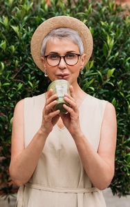 Stylish mature woman in a straw hat drinking a green smoothie in the park
