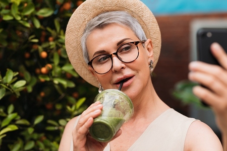 Senior woman taking a selfie outdoors while drinking a green smoothie  Portrait of a mature female in a small straw hat taking a selfie in the park