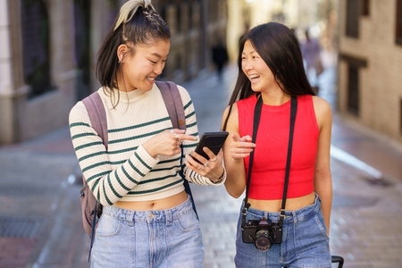 Cheerful Asian travelers using cellphone while walking on urban street