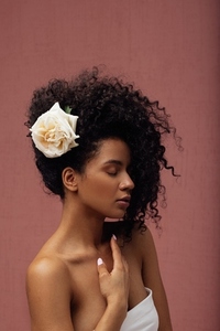 Young slim female with a white flower in her lush curly hair against a pink background