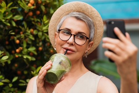 Stylish senior female drinking a smoothie and making selfie outdoors