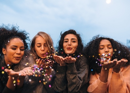 Group of four diverse females blowing colorful confetti from their hands at evening