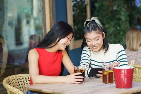 Cheerful Asian women sharing smartphone at table in cafe