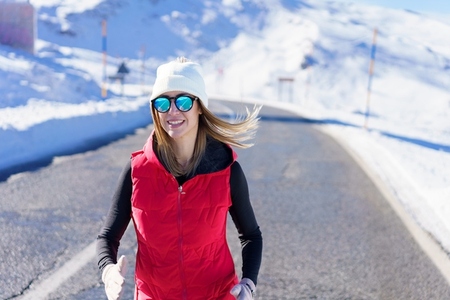 Happy young woman in warm vest running on road near snow
