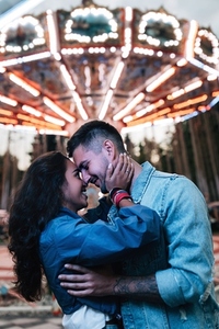 Young smiling couple in an amusement park hugging and kissing at night against a carousel with colorful lights
