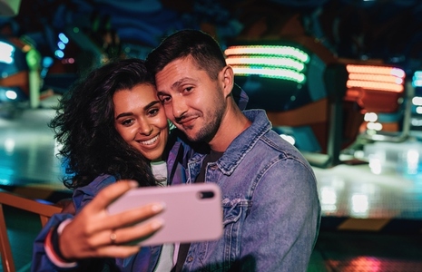 Young couple making selfie in amusement park against colorful lights