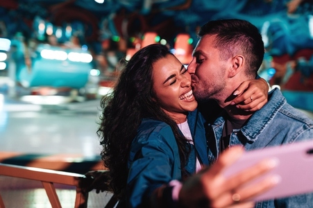 Boyfriend kissing his girlfriend while she is making selfie  Young couple during a summer festival