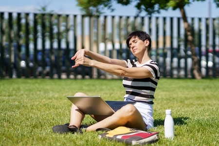 Female student stretching on green lawn in park and laptop with folder