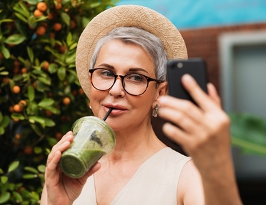 Aged woman in eyeglasses drinks smoothie and taking selfie outdoors